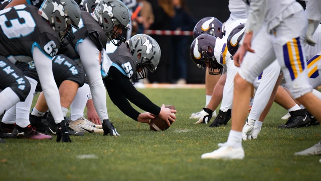 Zwei American-Football-Teams in Startposition gegenüber auf dem Spielfeld, ein Spieler hält den Ball zum Snap bereit