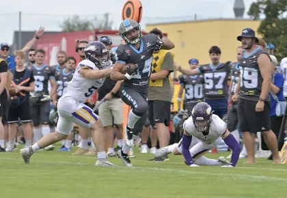 American-Football-Spieler in dunkler Uniform läuft mit Ball, während zwei Spieler in weißer Uniform versuchen, ihn zu stoppen, Zuschauer im Hintergrund